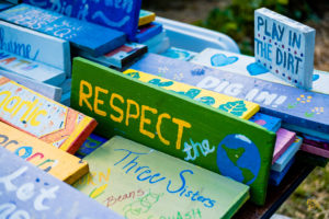 Colorful wooden signs with messages like "Respect the Earth," "Three Sisters," and "Play in the
