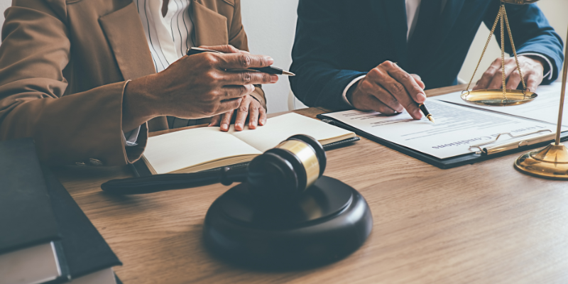 Two individuals discussing legal documents at a desk, with a gavel and scales of justice,