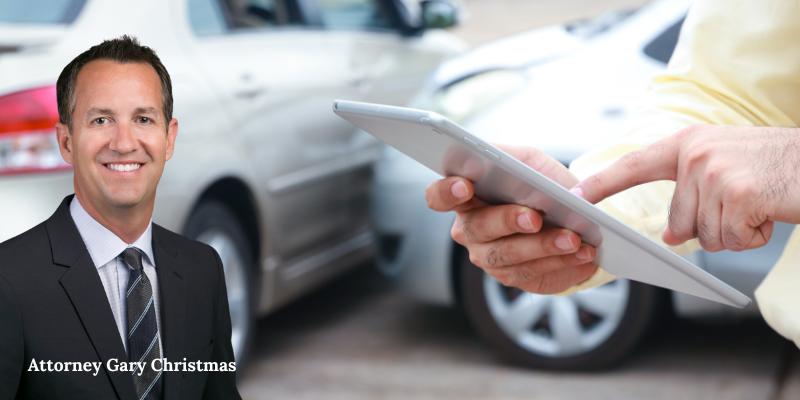 Attorney Gary Christmas standing beside a car accident scene, with a person using a tablet,
