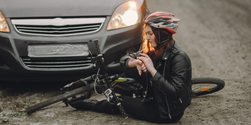 Bicyclist sitting on the ground after an accident, checking her wrist while a car is