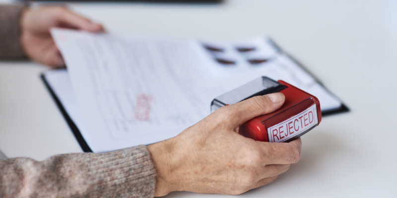 Person holding a red stamp labeled 'REJECTED' above a document, illustrating the challenges faced by